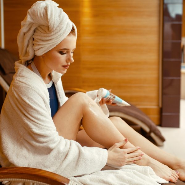 Sexy girl in bathrobe and towel on the head applying cream in spa chair, body care. Relaxation leisure, healthy lifestyle, attractive woman resting in armchair, beauty salon